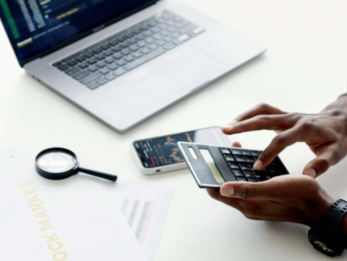 osoba unosi brojeve na digitronu, sa laptopom i telefonom sa dijagramima ispred na stolu